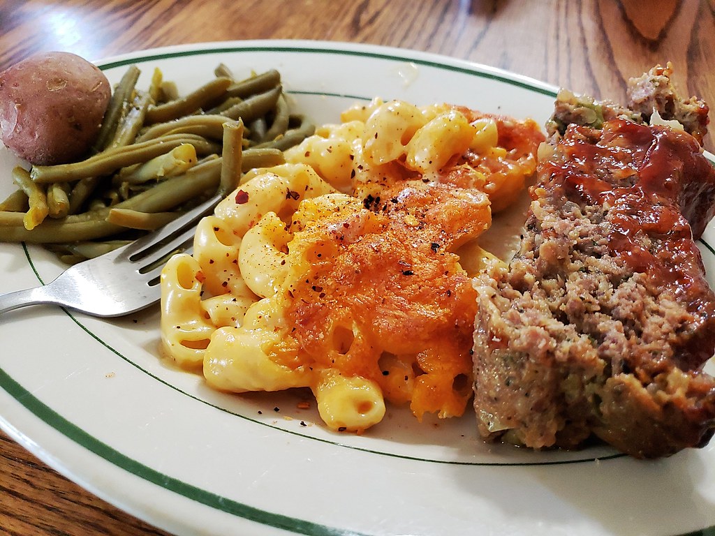 A homestyle dinner plate with meatloaf, macaroni and cheese, green beans, and a small potato.
