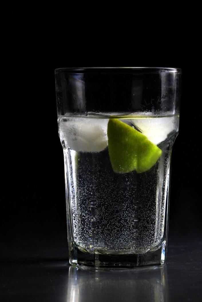 A tall highball glass filled with a clear sparkling drink, ice, and a lime wedge.